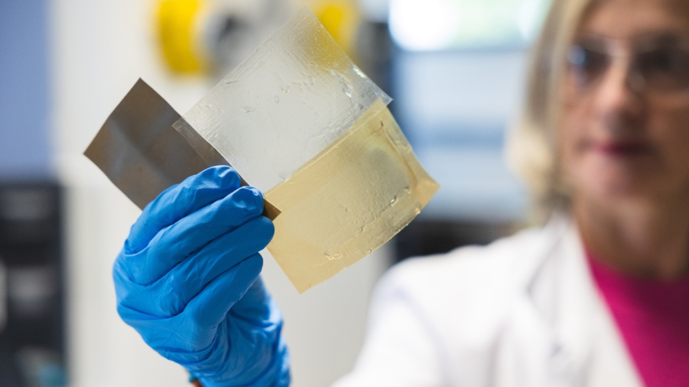A scientist wearing blue gloves holds up a translucent material sample in a laboratory setting.
