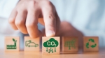 Hand selecting wooden block with CO2 reduction icon, surrounded by symbols of green factory, electric car, solar energy, and recycling, representing sustainable practices.